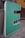 A three-dimensional letter 'E' in a vibrant mint green color, with a white border. It appears to be part of a larger sign or installation, resting on a concrete floor. The background shows a worn, industrial surface with patches of orange and gray, enhancing the urban, gritty atmosphere.
