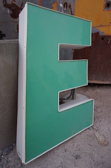 A three-dimensional letter 'E' in a vibrant mint green color, with a white border. It appears to be part of a larger sign or installation, resting on a concrete floor. The background shows a worn, industrial surface with patches of orange and gray, enhancing the urban, gritty atmosphere.