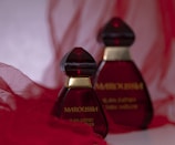 Two elegant perfume bottles with deep red glass and gold accents are placed against a soft, flowing red fabric background. The design is striking and sophisticated.