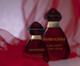 Two elegant perfume bottles with deep red glass and gold accents are placed against a soft, flowing red fabric background. The design is striking and sophisticated.