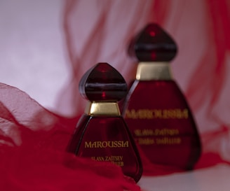Two elegant perfume bottles with deep red glass and gold accents are placed against a soft, flowing red fabric background. The design is striking and sophisticated.