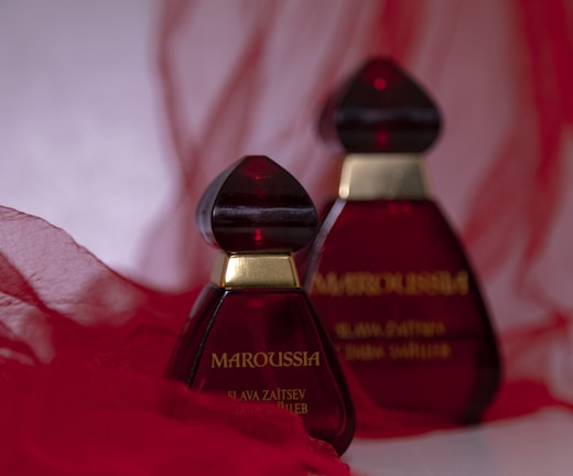 Two elegant perfume bottles with deep red glass and gold accents are placed against a soft, flowing red fabric background. The design is striking and sophisticated.