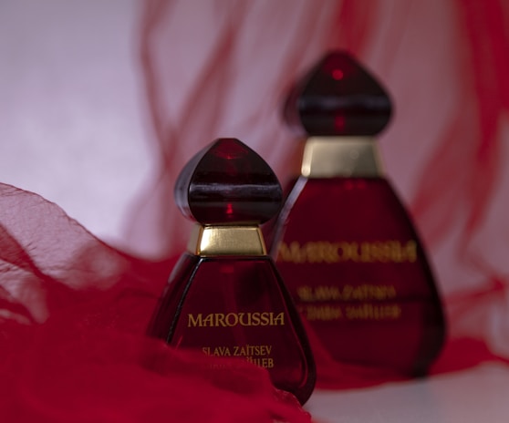 Two elegant perfume bottles with deep red glass and gold accents are placed against a soft, flowing red fabric background. The design is striking and sophisticated.