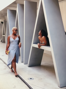 Two individuals are in a modern architectural setting with large, gray triangular structures. One person is walking and smiling, while the other is leaning playfully against one of the structures.
