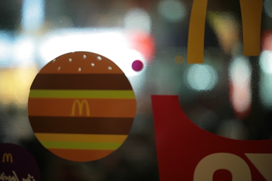 A blurred background with bright lights featuring a graphic of a burger and the McDonald's logo in front. The design is overlaid on a translucent surface with various shapes and colors.