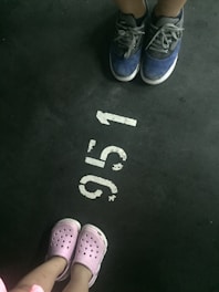 Two pairs of shoes are positioned on either side of a parking lot number. The top pair consists of blue sneakers with white soles, while the lower pair is light pink slip-on shoes with ventilation holes. The number '915' is painted in white on an asphalt ground.