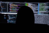 A dimly lit room with multiple computer screens displaying lines of code. The silhouette of a person sitting in front of the screens creates a mysterious and anonymous atmosphere. The code visible includes colorful syntax highlighting, typical of programming environments.