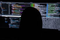A dimly lit room with multiple computer screens displaying lines of code. The silhouette of a person sitting in front of the screens creates a mysterious and anonymous atmosphere. The code visible includes colorful syntax highlighting, typical of programming environments.