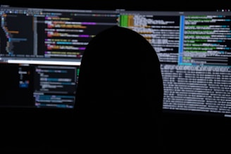 A dimly lit room with multiple computer screens displaying lines of code. The silhouette of a person sitting in front of the screens creates a mysterious and anonymous atmosphere. The code visible includes colorful syntax highlighting, typical of programming environments.