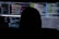 A dimly lit room with multiple computer screens displaying lines of code. The silhouette of a person sitting in front of the screens creates a mysterious and anonymous atmosphere. The code visible includes colorful syntax highlighting, typical of programming environments.