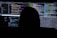 A dimly lit room with multiple computer screens displaying lines of code. The silhouette of a person sitting in front of the screens creates a mysterious and anonymous atmosphere. The code visible includes colorful syntax highlighting, typical of programming environments.