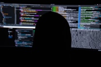 A dimly lit room with multiple computer screens displaying lines of code. The silhouette of a person sitting in front of the screens creates a mysterious and anonymous atmosphere. The code visible includes colorful syntax highlighting, typical of programming environments.