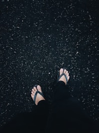 person in black flip flops and black pants standing on black asphalt floor
