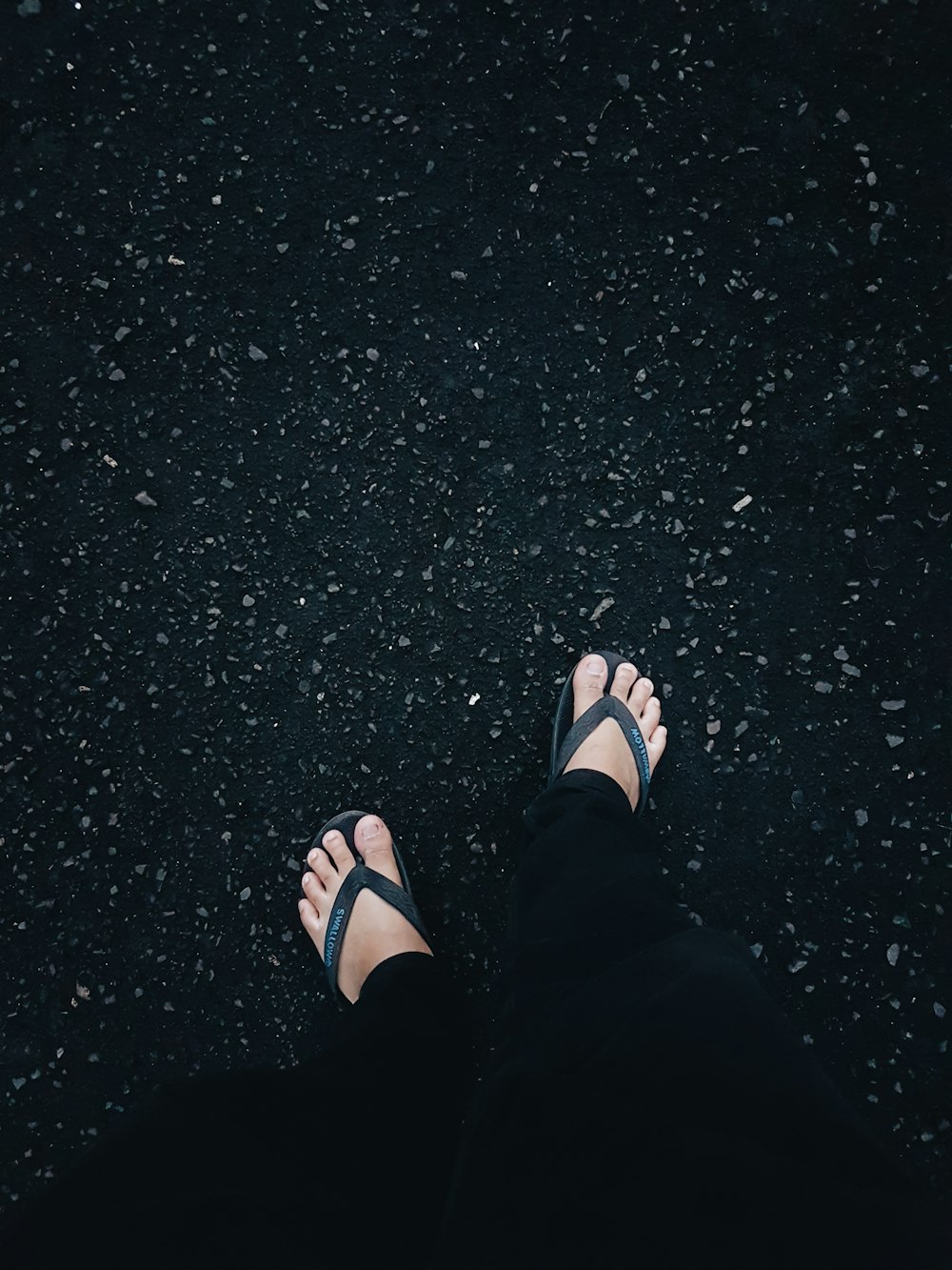person in black flip flops and black pants standing on black asphalt floor