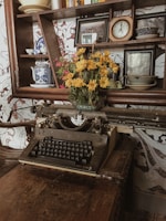 An antique typewriter sits on a worn wooden table, surrounded by vintage items. Above the typewriter, a shelf holds decorative plates, a clock, a framed picture, and a vibrant bouquet of yellow flowers in a glass vase, adding a touch of color to the rustic scene.