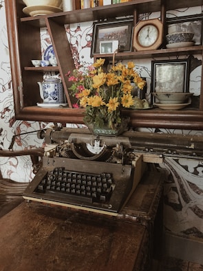 An antique typewriter sits on a worn wooden table, surrounded by vintage items. Above the typewriter, a shelf holds decorative plates, a clock, a framed picture, and a vibrant bouquet of yellow flowers in a glass vase, adding a touch of color to the rustic scene.
