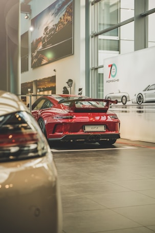A red sports car with GT3 branding is displayed in a modern showroom. The showroom features large glass windows and several large posters on the walls, depicting similar vehicles, reinforcing the high-performance theme. The environment is spacious and well-lit, emphasizing the sleek design of the car.
