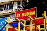 A vibrant sign with bold yellow text on a red background reads 'BY MUMBAI.' It is mounted on a wooden structure with colorful panels beneath it. The background shows part of a building with windows and some greenery.