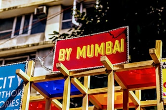 A vibrant sign with bold yellow text on a red background reads 'BY MUMBAI.' It is mounted on a wooden structure with colorful panels beneath it. The background shows part of a building with windows and some greenery.