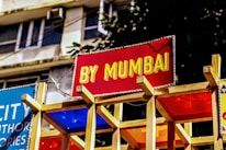 A vibrant sign with bold yellow text on a red background reads 'BY MUMBAI.' It is mounted on a wooden structure with colorful panels beneath it. The background shows part of a building with windows and some greenery.