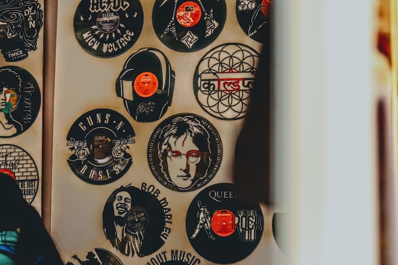 An array of vinyl records on a wall, each featuring the likeness or logo of famous musicians and bands such as Bob Marley, Guns N' Roses, Queen, and John Lennon. Some records have a central image cut out or printed, resembling popular album art or artist portraits, with vibrant splashes of color and artistic detail.