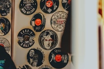An array of vinyl records on a wall, each featuring the likeness or logo of famous musicians and bands such as Bob Marley, Guns N' Roses, Queen, and John Lennon. Some records have a central image cut out or printed, resembling popular album art or artist portraits, with vibrant splashes of color and artistic detail.