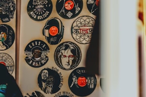 An array of vinyl records on a wall, each featuring the likeness or logo of famous musicians and bands such as Bob Marley, Guns N' Roses, Queen, and John Lennon. Some records have a central image cut out or printed, resembling popular album art or artist portraits, with vibrant splashes of color and artistic detail.