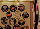 A wall decorated with vinyl records featuring various iconic rock bands and musicians. Each record displays band logos and artwork, including bands like AC/DC, Metallica, Guns N' Roses, Led Zeppelin, and The Beatles. The display is illuminated by a ceiling light, creating a warm ambiance. T-shirts hang on the right side.
