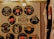 A wall decorated with vinyl records featuring various iconic rock bands and musicians. Each record displays band logos and artwork, including bands like AC/DC, Metallica, Guns N' Roses, Led Zeppelin, and The Beatles. The display is illuminated by a ceiling light, creating a warm ambiance. T-shirts hang on the right side.