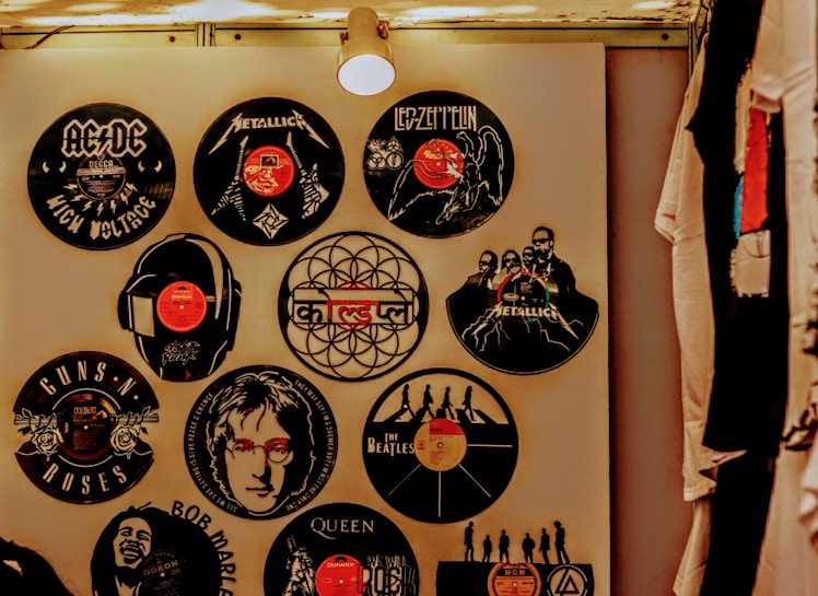 A wall decorated with vinyl records featuring various iconic rock bands and musicians. Each record displays band logos and artwork, including bands like AC/DC, Metallica, Guns N' Roses, Led Zeppelin, and The Beatles. The display is illuminated by a ceiling light, creating a warm ambiance. T-shirts hang on the right side.
