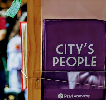 A wooden frame supporting a purple book or banner with the words 'City's People' and 'Pearl Academy' printed on it. The background features blurred colors and shapes, suggesting a busy or crowded environment.