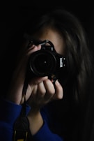 A person holding a camera up to their face, capturing an image. The scene is dimly lit, focusing on the camera and the person's hands. The background is dark, emphasizing the photographer's concentration.