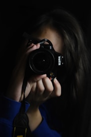 A person holding a camera up to their face, capturing an image. The scene is dimly lit, focusing on the camera and the person's hands. The background is dark, emphasizing the photographer's concentration.