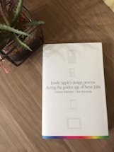 A hardcover book with a white cover is placed on a wooden surface. The book is titled 'Inside Apple's design process during the golden age of Steve Jobs' by Ken Kocienda. The cover features minimalist icons and a colorful strip at the bottom. Next to the book, there is a plant in a wicker basket.