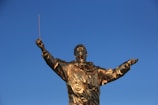 A golden statue of a person wearing a space suit, holding a baton in an elevated position against a clear blue sky.