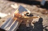 A block of wood rests on a tree stump with curly wood shavings artistically arranged on top. The sunlight casts long shadows, highlighting the texture and grooves of the wood.