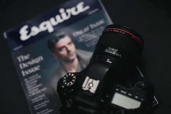 A DSLR camera with a 35mm lens rests on a magazine. The magazine has a blurred image of a person on the cover and the text 'Esquire' prominently featured. The camera's lens has a red ring and is positioned on top of the publication, suggesting a photography theme.