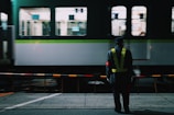 A railway station scene at night with a guard standing in front of a moving train. The guard wears a reflective vest and cap, and stands behind a safety barrier. The train in the background is green and white, with blurred windows indicating motion.