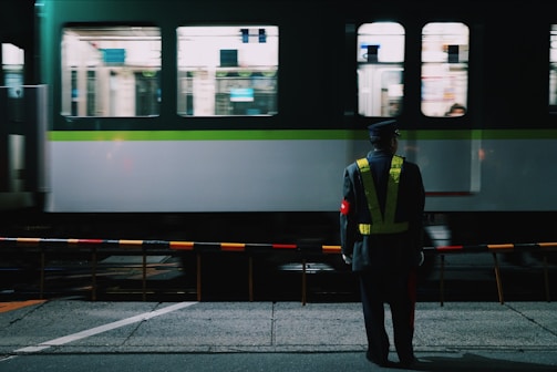 A railway station scene at night with a guard standing in front of a moving train. The guard wears a reflective vest and cap, and stands behind a safety barrier. The train in the background is green and white, with blurred windows indicating motion.