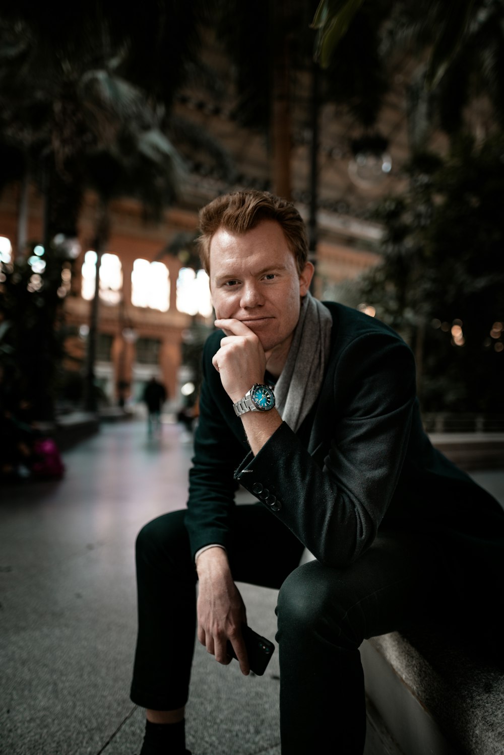 man sitting on pavement wearing leather jacket and dress pants holding smartphone near trees with the distance of building during night time