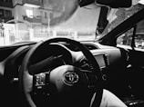 The interior of a car featuring a steering wheel, dashboard, and infotainment system. The windshield displays a view of residential buildings outside, seen through a slightly foggy glass.