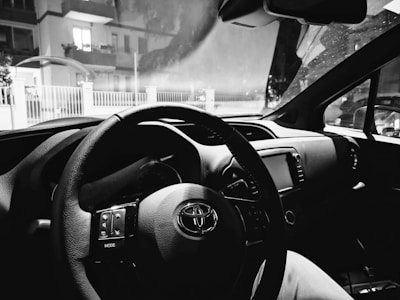 The interior of a car featuring a steering wheel, dashboard, and infotainment system. The windshield displays a view of residential buildings outside, seen through a slightly foggy glass.