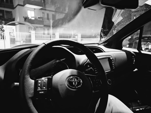 The interior of a car featuring a steering wheel, dashboard, and infotainment system. The windshield displays a view of residential buildings outside, seen through a slightly foggy glass.