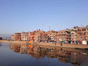 A row of traditional brick buildings lines a riverside with their reflections visible in the calm water. The architecture features multiple stories and a classic design, with various small shops and antennas visible on rooftops. People are walking along the riverside, and there is a bright orange object, possibly a tent or awning, near the buildings.