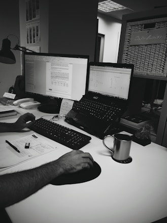 A monochrome image of an office desk setup with two computer monitors displaying documents. One monitor is on a stand, while the other is a laptop. A person is using a keyboard and mouse. There are papers, a pen, and a phone on the desk. A metal mug sits on a coaster nearby. In the background, a partially visible basket with a label is mounted on a wall.
