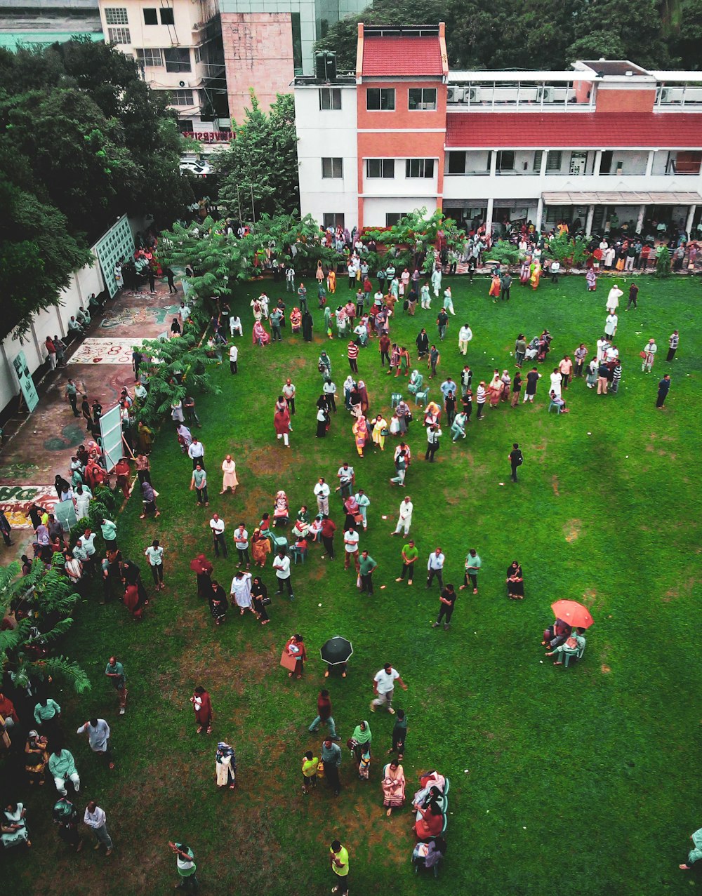 Fotografía aérea de personas caminando en un campo verde cerca de un edificio de 3 pisos de concreto blanco y rojo durante el día