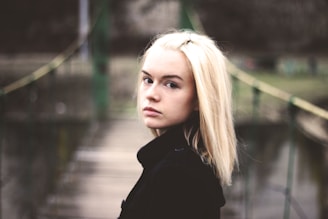 shallow focus photo of young blond girl in black coat standing on a bridge