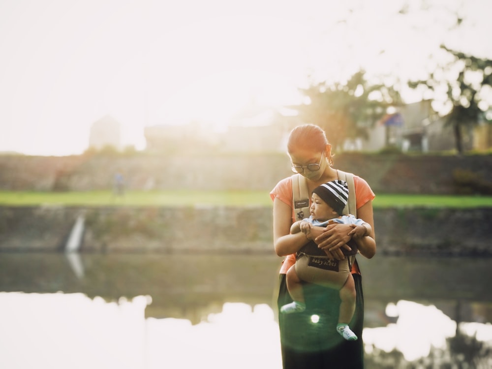 woman carrying a baby with brown carrier while standing near body of water