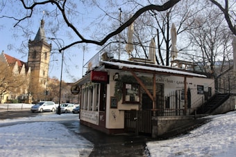 A small cafe or bar situated at the corner of a quiet street during winter. The building is surrounded by bare trees and has outdoor seating with umbrellas. Snow partially covers the ground, and there are several cars parked along the street. In the background, there's a historic-looking tower, possibly part of a church or similar structure.
