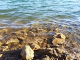 Clear water gently washes over a collection of stones and rocks, revealing their textures and colors beneath the surface. The sunlight creates a shimmering effect on the water, adding a sense of tranquility.
